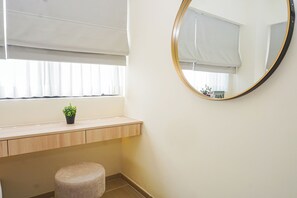 A room with a wooden desk, a round mirror, a small stool, and a window with blinds. - Cozy And Functional 3Br At Apartment Meikarta (Cikarang)