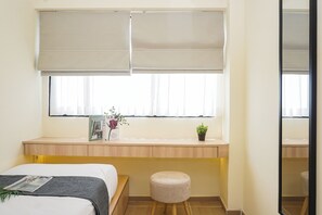A bedroom with a bed, a bedside table with a book and a potted plant, a window with blinds, and a small stool. - Cozy And Functional 3Br At Apartment Meikarta (Cikarang)