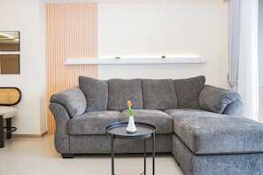 A modern living room with a grey sectional sofa, a small round table, and a white shelf above the sofa. - Cozy And Functional 3Br At Apartment Meikarta (Cikarang)