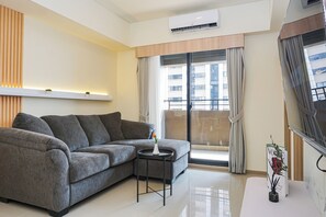 A modern living room with a grey sofa, a small round table, and a window with curtains. - Cozy And Functional 3Br At Apartment Meikarta (Cikarang)