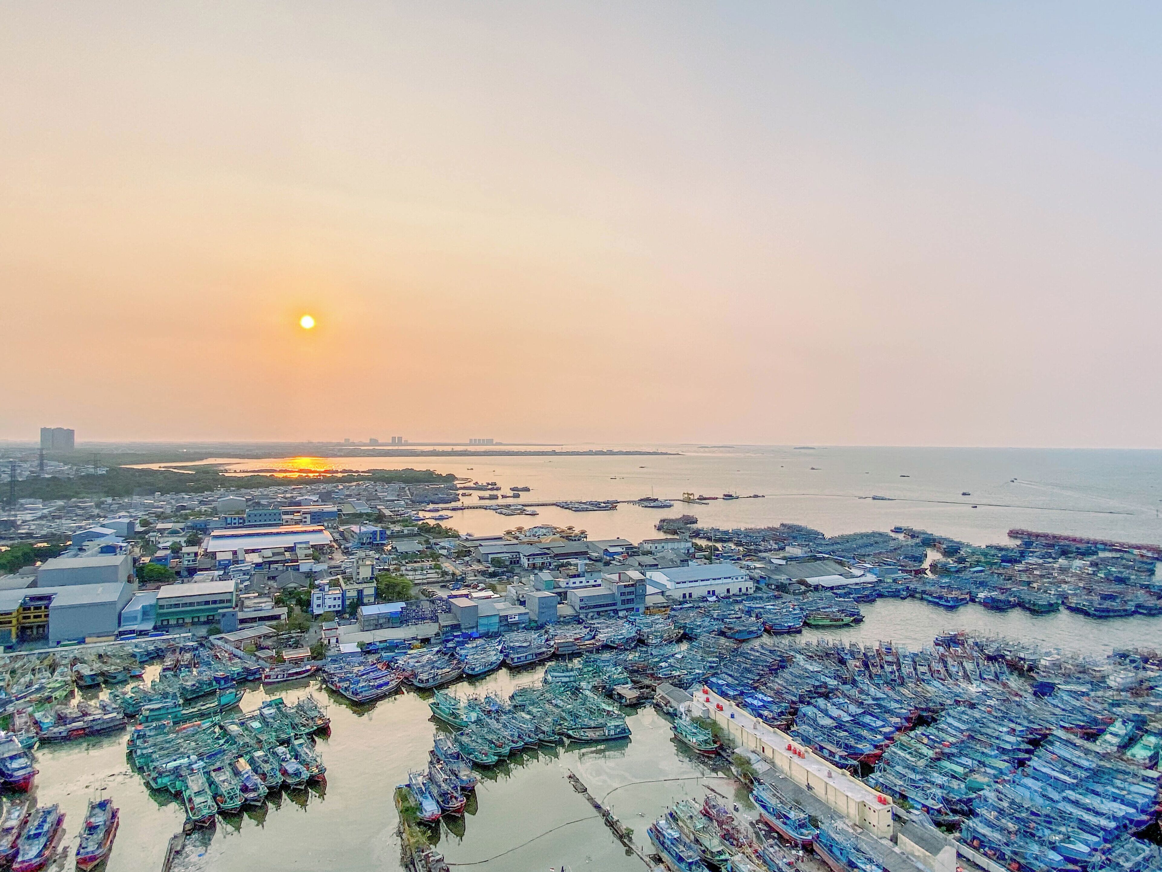 Eine Küstenstadt mit einer dichten Ansammlung von Booten im Vordergrund und freier Sicht auf den Sonnenuntergang über dem Wasser.