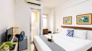 A hotel room with a bed, a TV, a fan, a small table with a vase of flowers, and a picture on the wall. - Nice And Warm Studio Room At Apartment Green Bay Pluit (Jakarta)