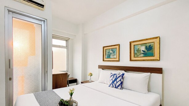 A hotel room with a bed, a desk, a chair, a window, and two framed pictures on the wall. - Nice And Warm Studio Room At Apartment Green Bay Pluit (Jakarta)