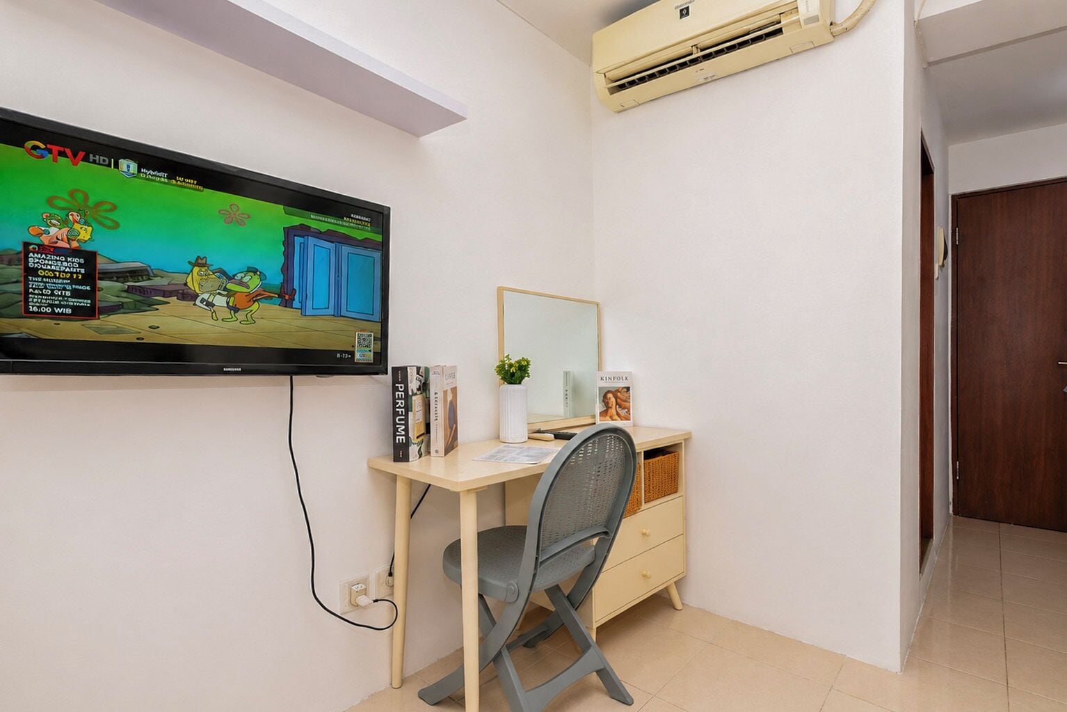 Ein Zimmer mit an der Wand befestigtem Fernseher, einem Schreibtisch mit Stuhl, einem Spiegel und einer kleinen Pflanze.