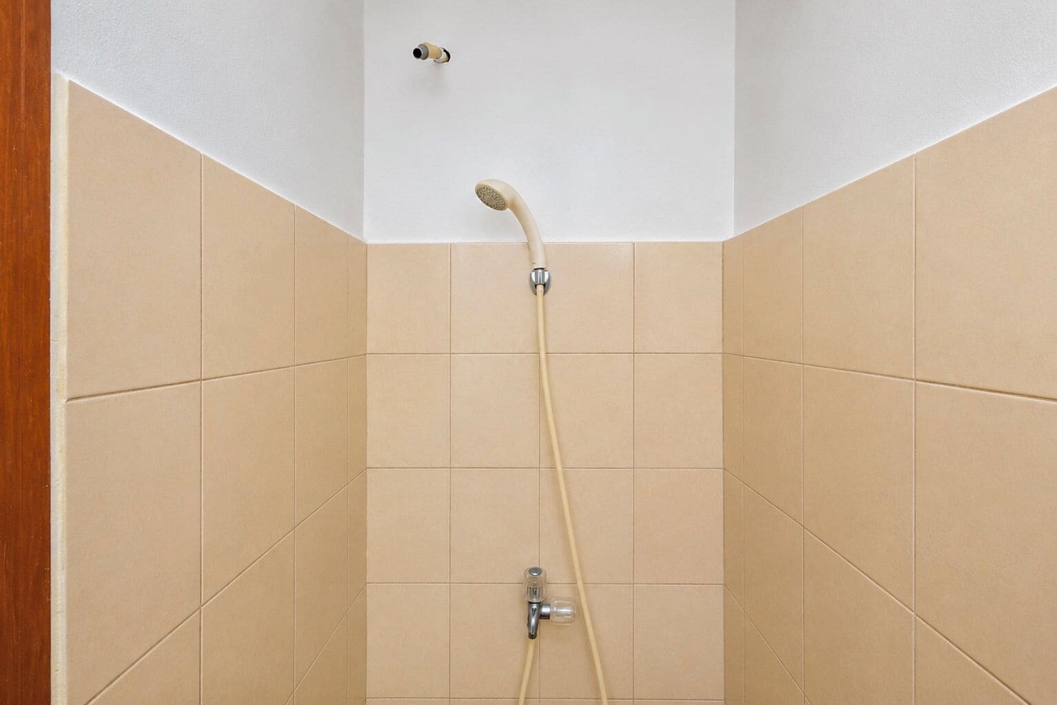 Ducha con cabezal de ducha de mano y grifo empotrado en la pared, en una esquina de una habitación con paredes de azulejos color beige.