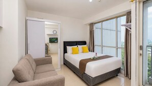 A hotel room with a bed, a sofa, a television, and a view of the city through large windows. - Minimalist And Homey Studio At Apartment Tifolia (Jakarta)