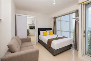 Quarto de hotel com cama, sofá, televisão e vista da cidade através de grandes janelas.