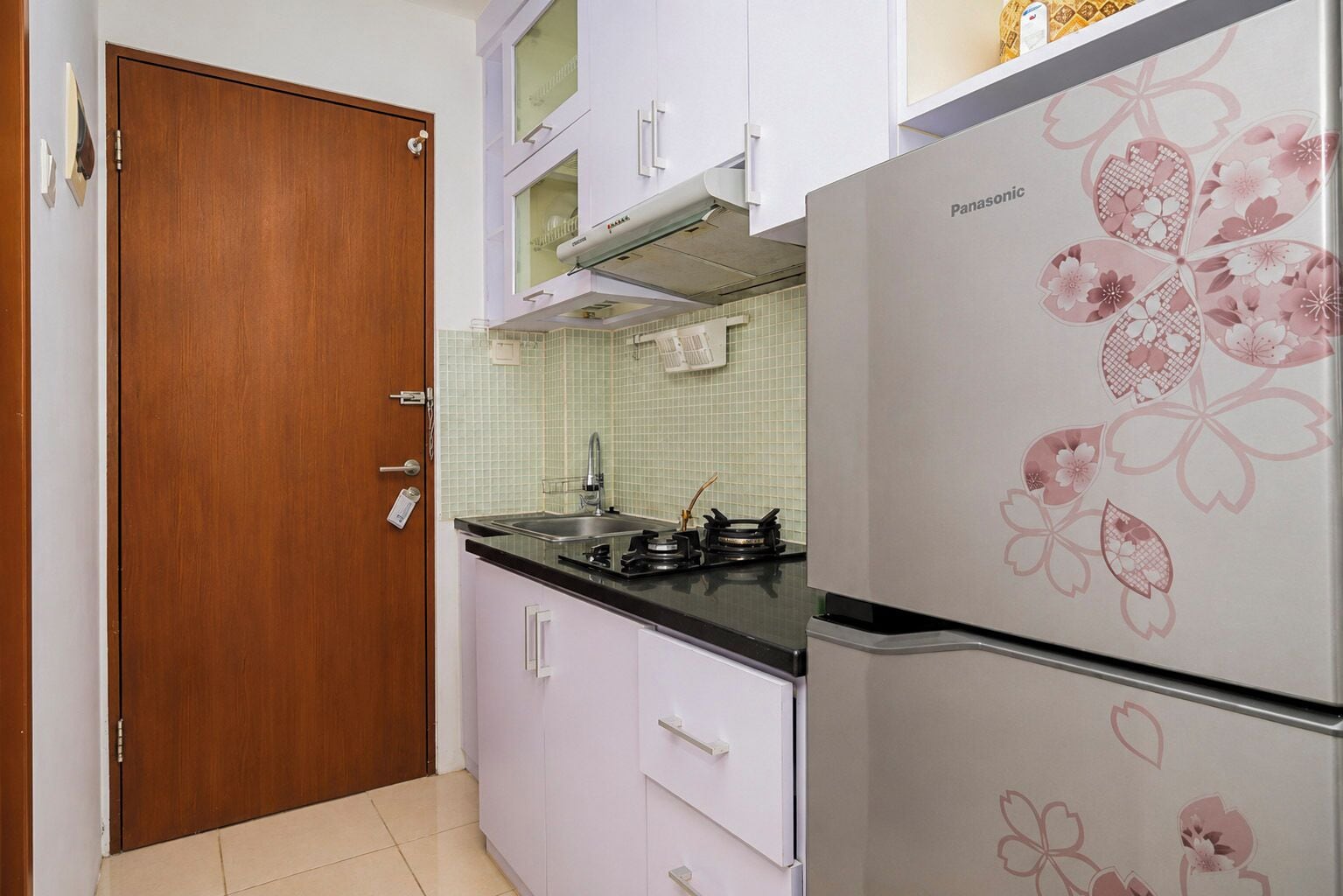 Een keuken met een houten deur, witte kastjes, een koelkast met bloemendecor en een betegelde achterwand.