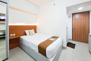A hotel room with a bed, a wooden headboard, a bedside table, a TV, and a door. - Functional Simple Studio Room At Apartment Cisauk Point (Cisauk)