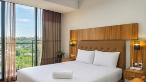 A hotel room with a large bed, wooden headboard, two bedside lamps, a view of greenery outside, and a potted plant on the nightstand. - Modern Breezy Studio At Apartment Tamansari Skylounge Balikpapan (Balikpapan)