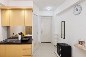 A compact kitchen with a sink, cabinets, and a countertop. There is a door leading to another room, a clock on the wall, and a shelf with a framed picture. - Best Studio Apartment Green Pramuka City (Jakarta)
