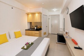 Quarto de hotel com cama, pequeno espaço de cozinha, televisão e mesa de cabeceira.