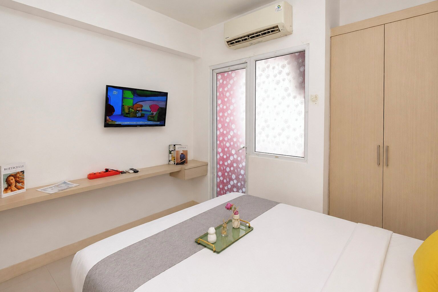 A hotel room with a bed, a television mounted on the wall, a wardrobe, and a window with a floral curtain.
