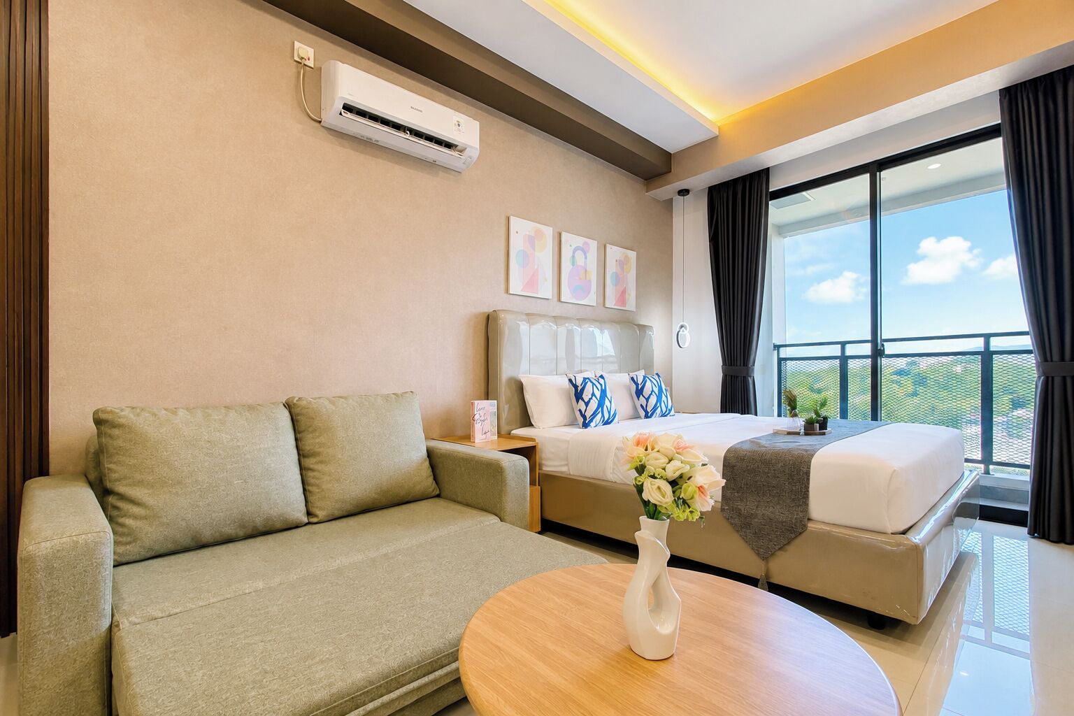 A hotel room with a sofa, a bed with decorative pillows, a bedside table with a vase, and a large window with a view.