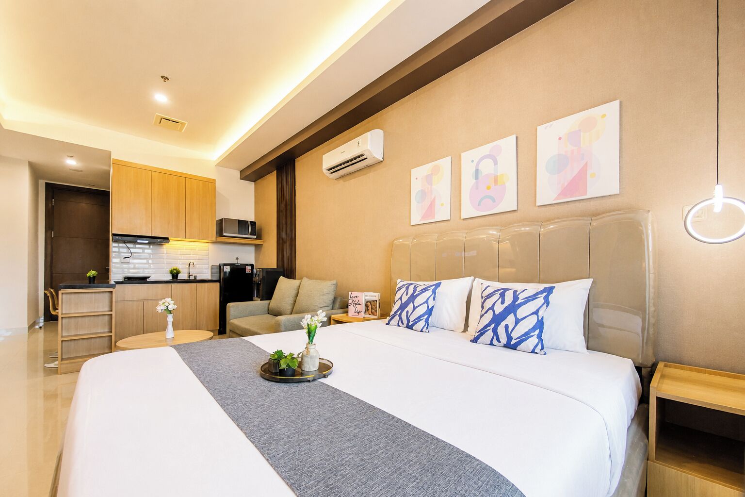 A modern hotel room with a large bed, a bedside table, a nightstand, a sofa, a kitchen area, and a wall with abstract art.