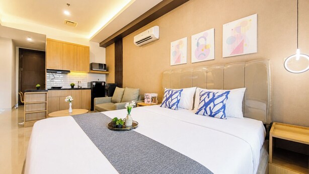 A modern hotel room with a large bed, a bedside table, a nightstand, a sofa, a kitchen area, and a wall with abstract art. - Cozy Living Studio At Apartment Saffron Noble (Bogor)