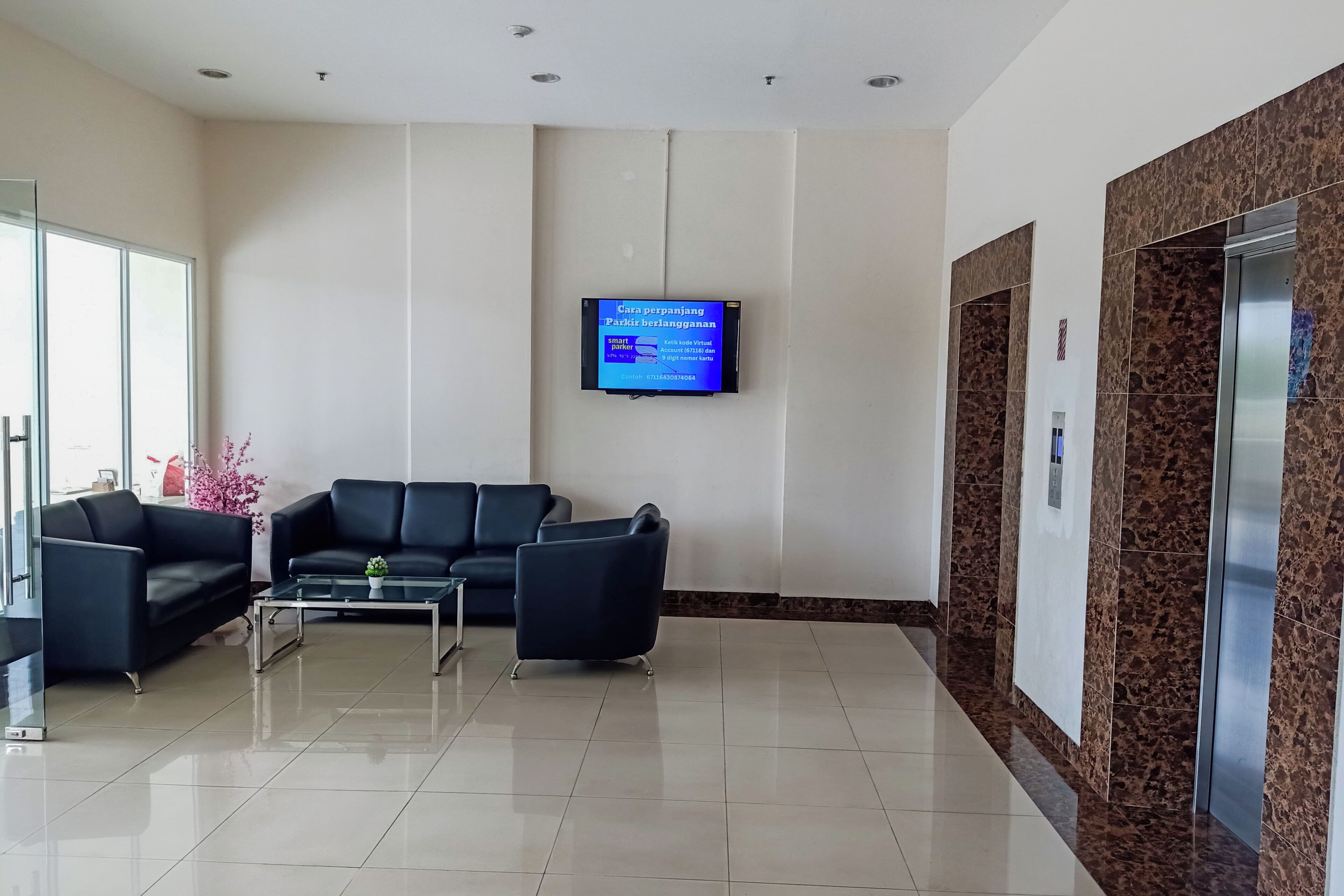 Uma área de recepção com um sofá de couro preto, uma mesa de centro de vidro e uma televisão de tela plana na parede exibindo uma tela com informações sobre estacionamento.