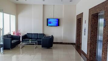 Uma área de recepção com um sofá de couro preto, uma mesa de centro de vidro e uma televisão de tela plana na parede exibindo uma tela com informações sobre estacionamento.