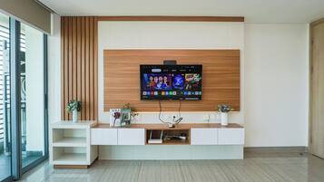 A modern living room with a flat-screen TV mounted on a wooden wall, a white entertainment unit below, and a glass door leading to a balcony.