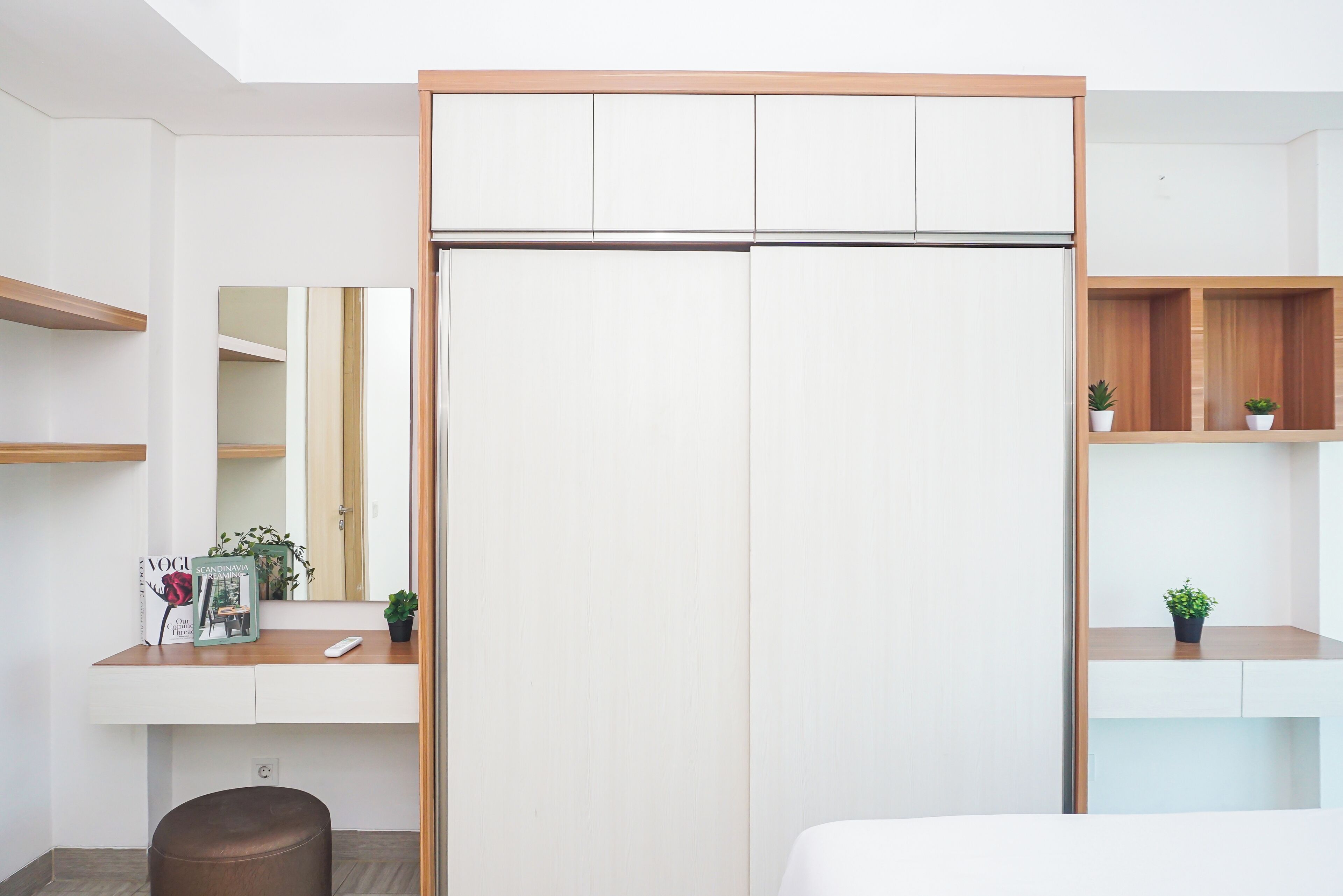 Un dormitorio moderno con un armario, una mesita de noche con un libro y un control remoto, un pequeño taburete y una planta en maceta.