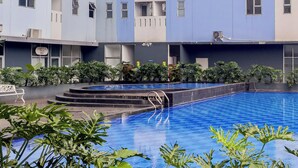 A swimming pool with steps leading into it, surrounded by greenery and a multi-story building with balconies. - Nice And Tidy 1Br At Apartment Urbantown Serpong (South Tangerang)