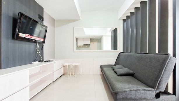 A modern living room with a grey sofa, a flat-screen TV mounted on the wall, a small white table, and a large mirror reflecting the room. - Nice And Tidy 1Br At Apartment Urbantown Serpong (South Tangerang)