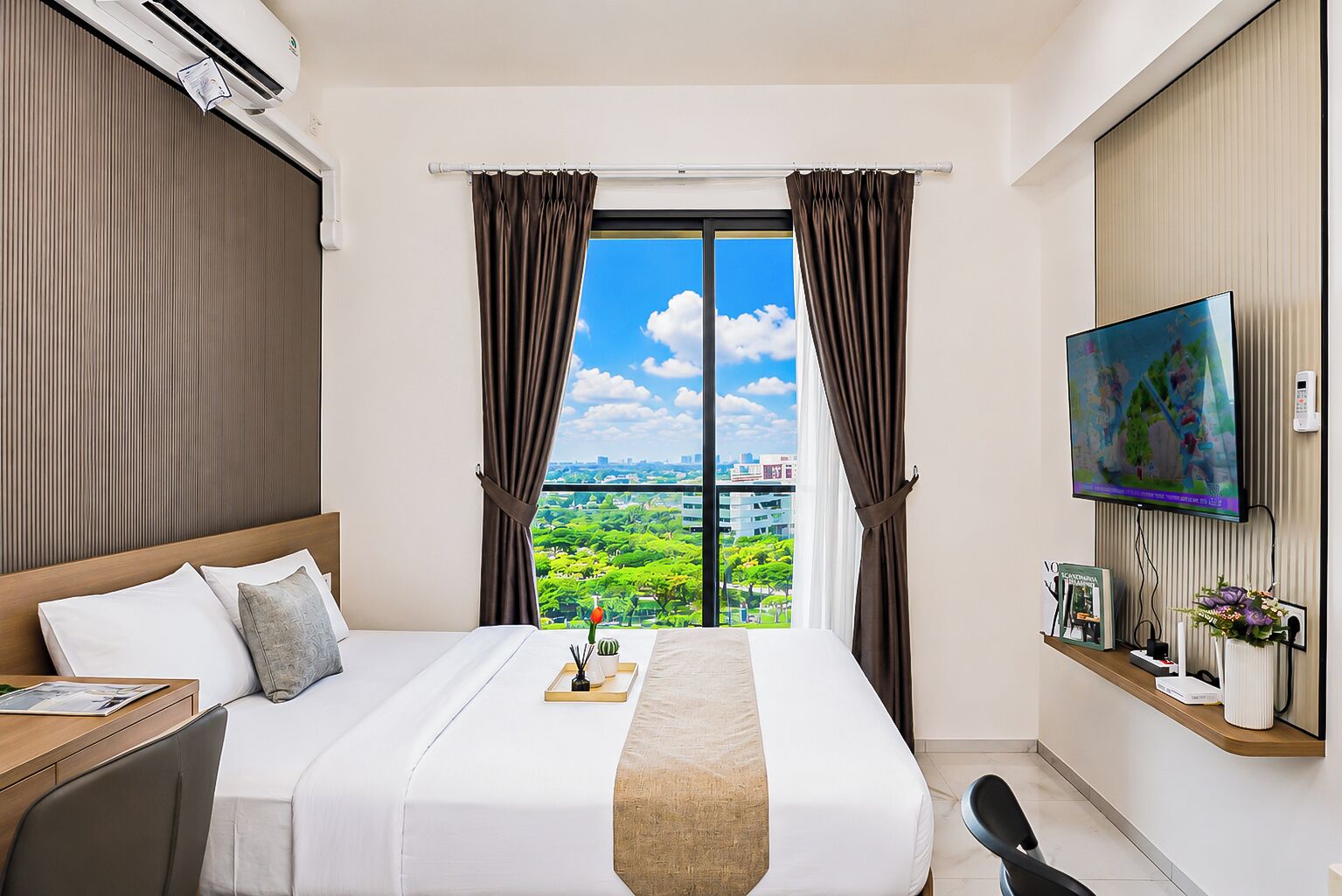 Ein Hotelzimmer mit einem großen Bett, einem Schreibtisch, einem Sessel, einem Fernseher und Blick auf eine Stadtlandschaft durch ein Fenster.