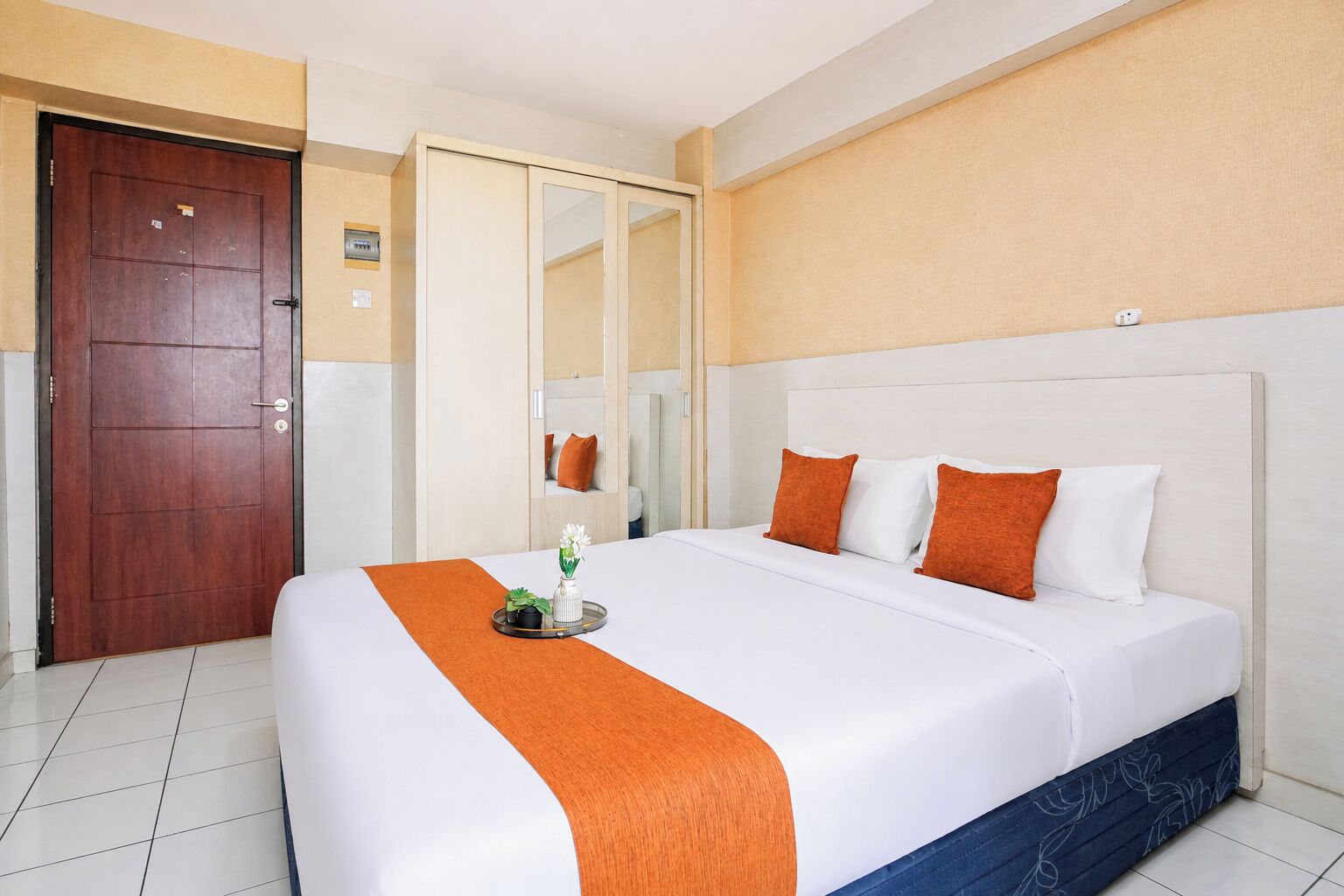 Habitación de hotel con una cama grande, una puerta de madera, un espejo y una pared blanca.