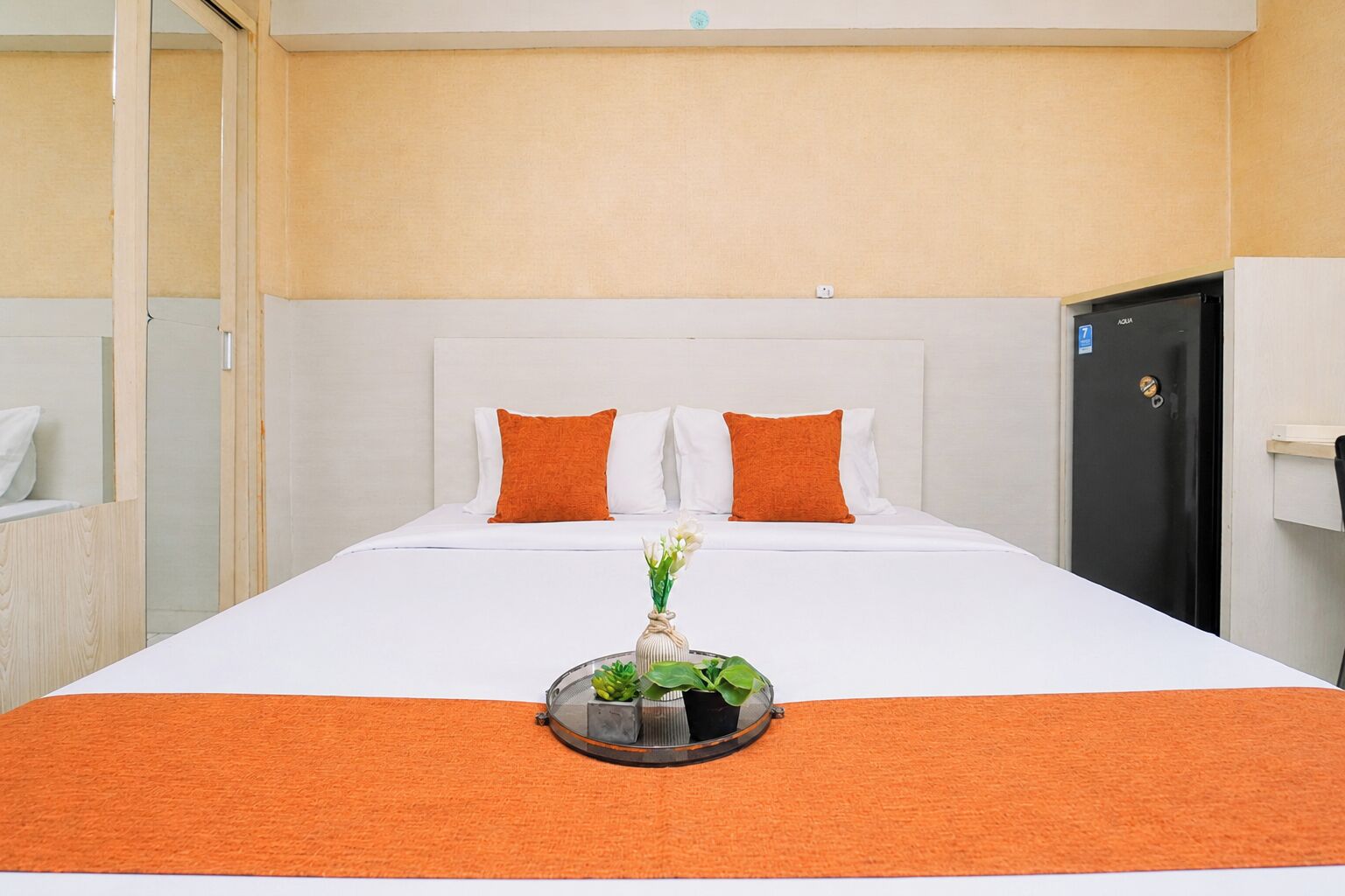 A hotel room with a bed, two pillows, a nightstand, a small table with a vase and flowers, and a door with a lock.
