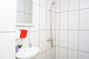 Ein Badezimmer mit weißen Fliesen, einem weißen Waschbecken, einer roten Mülltonne und einer Dusche mit Duschbrause.