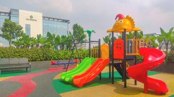A playground with slides and a climbing structure, surrounded by greenery and a building in the background.