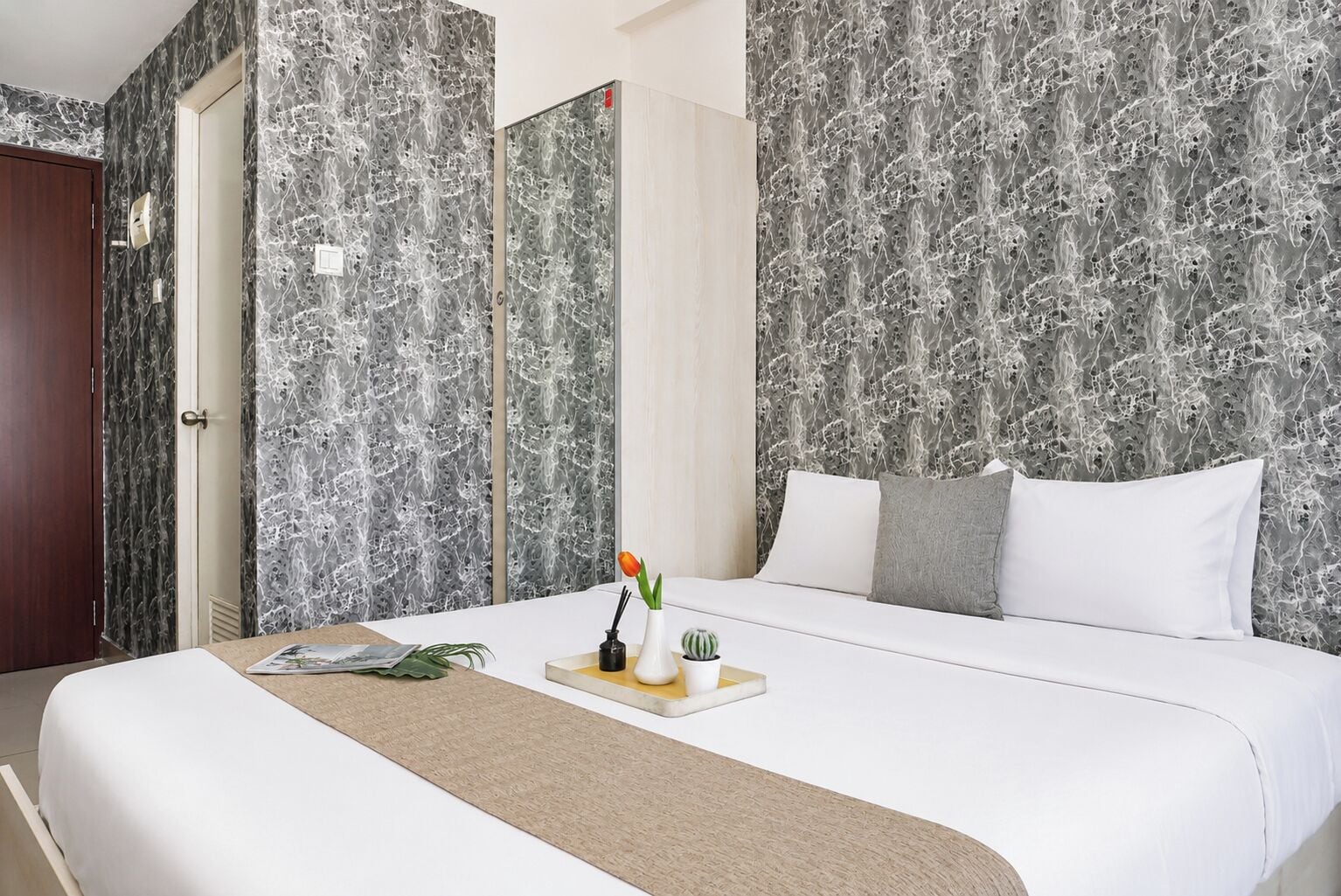 Habitación de hotel con una cama grande, una mesita de noche con un florero y una planta, y una puerta con interruptor de luz.
