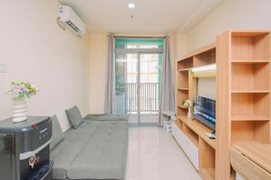 A modern living room with a grey sofa, a flat-screen TV on a wooden stand, and a wall-mounted air conditioner. - Enjoy Living 1Br Apartment At Pejaten Park Residence (Jakarta)