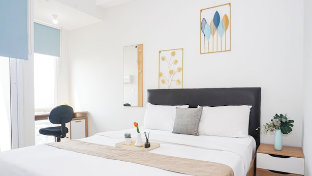 Quarto de hotel com cama, escrivaninha, cadeira, mesa de cabeceira, um vaso com flores e decoração na parede.
