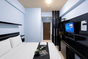 A hotel room with a bed, a TV on the wall, a small table with a vase and flowers, and a door leading to another room. - Chic Minimalist Studio At Apartment Suites @Metro (Bandung)