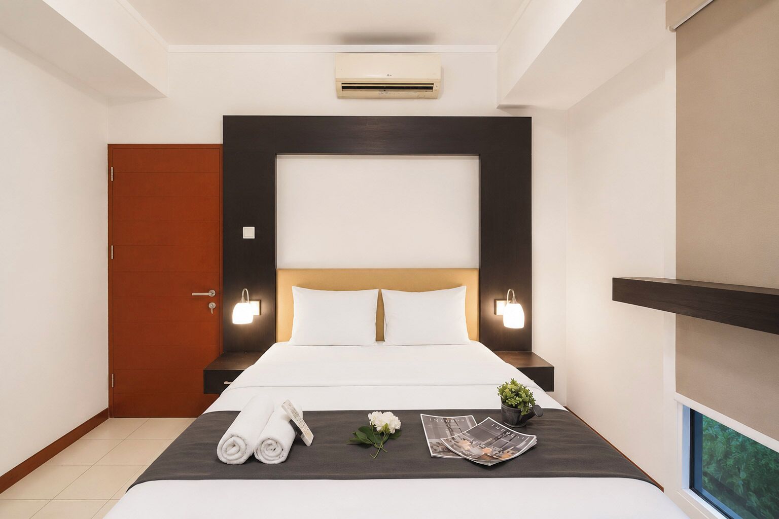 A hotel room with a bed, bedside tables, an air conditioning unit, a door, and a window.