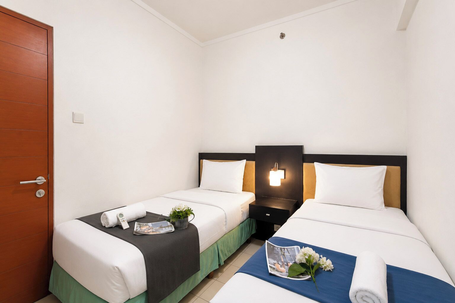 Ein Hotelzimmer mit zwei Betten, einer Holztür, einem Nachttisch mit Lampe und einem kleinen Tisch mit Zeitschrift und Blumen.