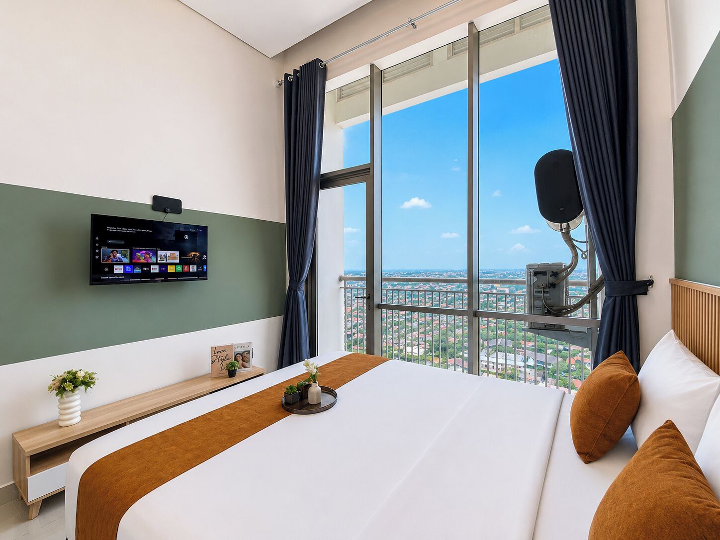 Habitación de hotel con un ventanal grande, un televisor de pantalla plana montado en la pared, una cama con almohadas marrones y una mesita de noche pequeña con una planta.