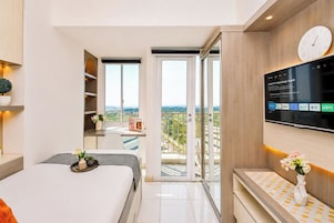 A modern hotel room with a bed, a desk, a TV mounted on the wall, and a balcony with a city view.