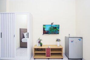 Hotellihuone, jossa on sänky, seinälle asennettu televisio, pieni jääkaappi ja puinen kaappi, jossa on maljakko ja kirja.