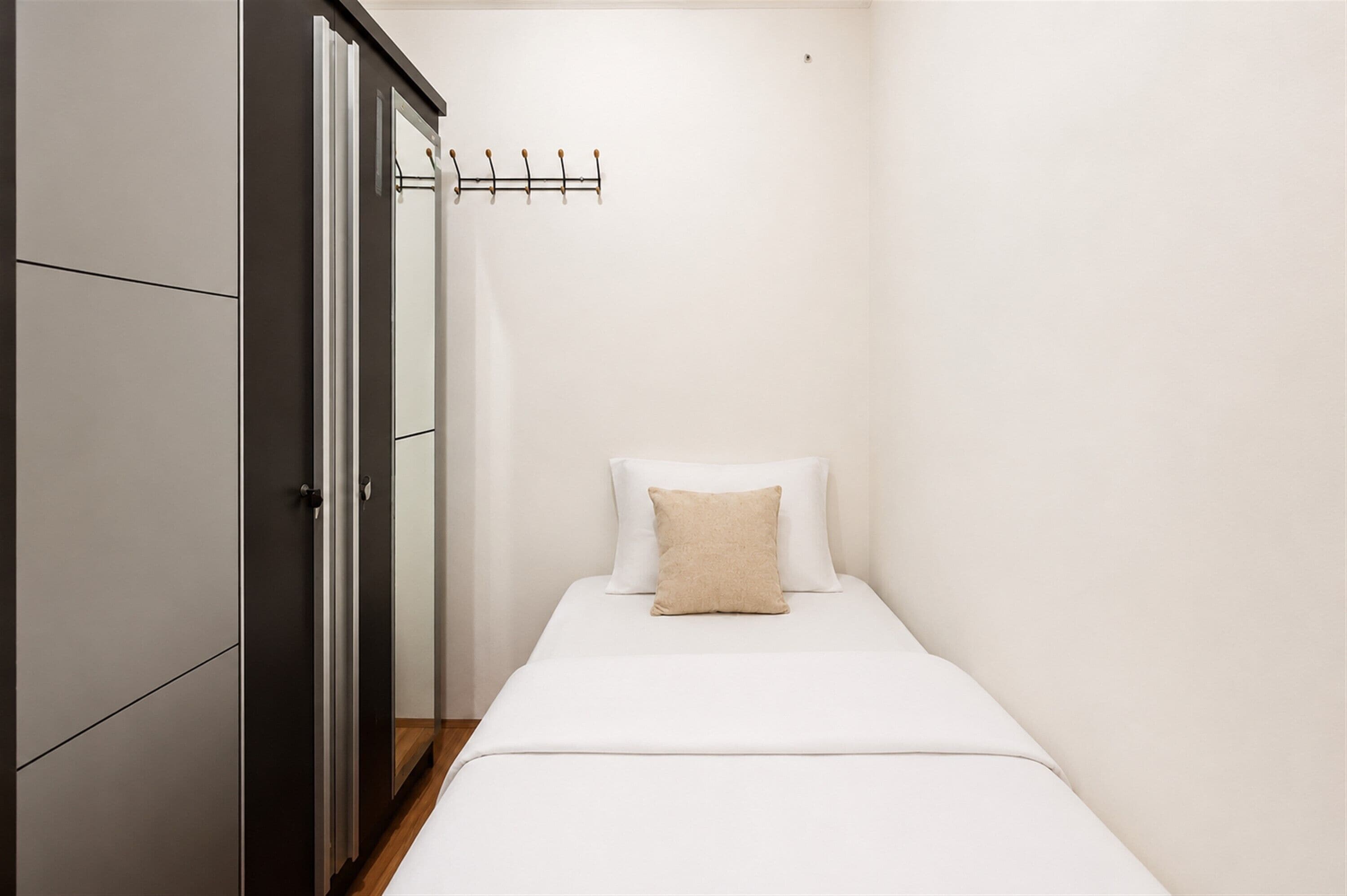 Een kleine kamer met een eenpersoonsbed, een garderobe en een kapstok.