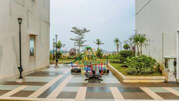 Un parque infantil con una estructura para escalar y un tobogán, rodeado de un área pavimentada con un farol y vegetación.