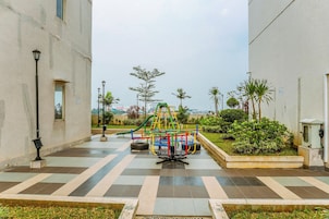 A playground with a climbing structure and a slide, surrounded by a paved area with a lamp post and greenery.