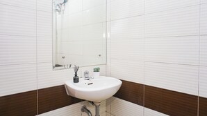 Een badkamer met witte betegelde wanden, een witte wastafel, een spiegel, een kraan, een potplant en een klein vaasje met rietstengels.