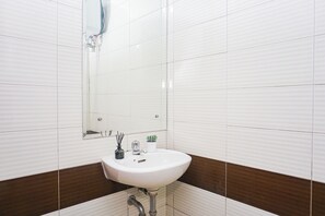 A bathroom with white tiled walls, a white sink, a mirror, a faucet, a potted plant, and a small vase with reeds. - Comfortable Living In Studio Room At Apartment Margonda Residence 3 (Depok)