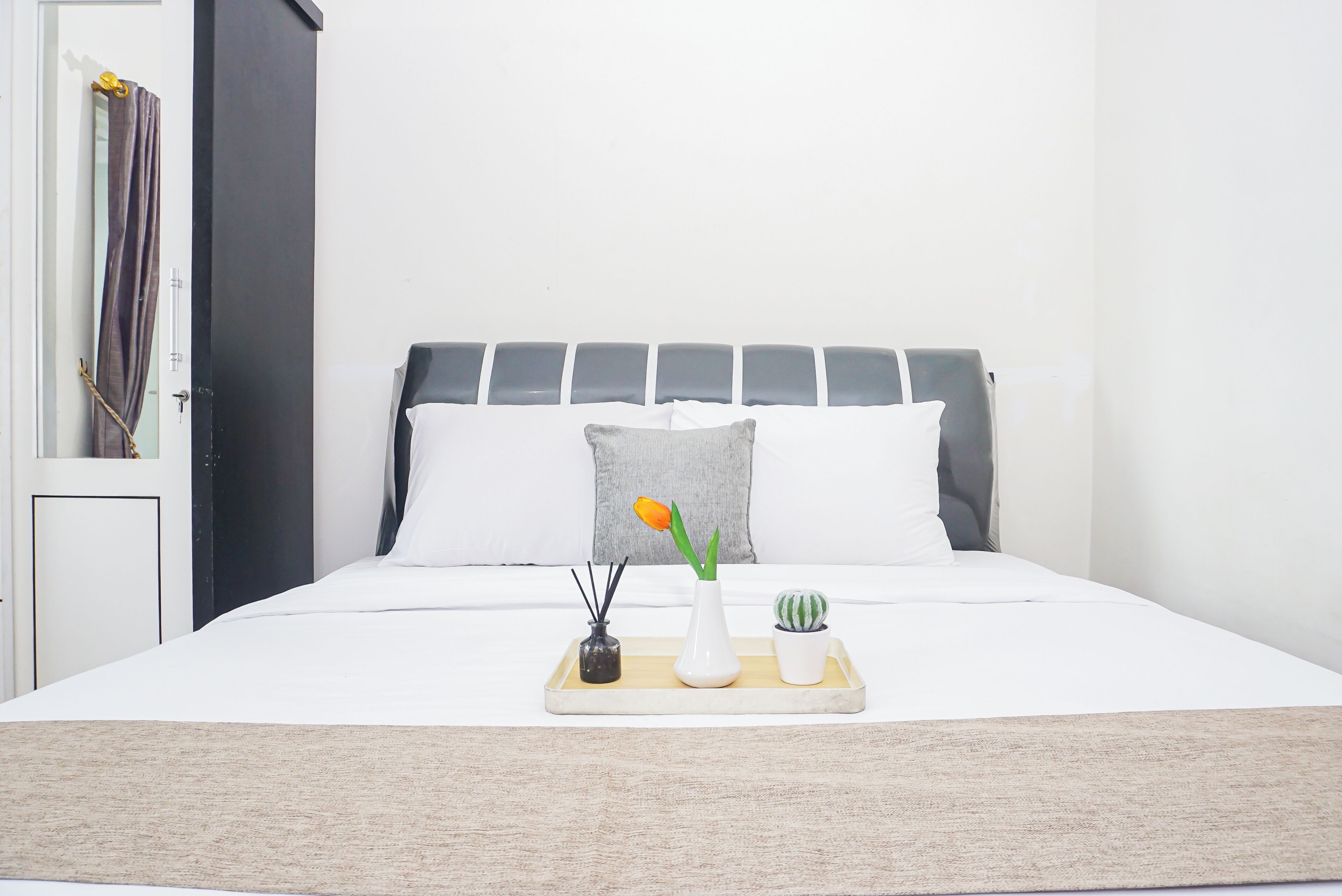 A bed with a white mattress, grey headboard, and white pillows. A tray with a vase, a small plant, and a candle holder is placed on the bed.