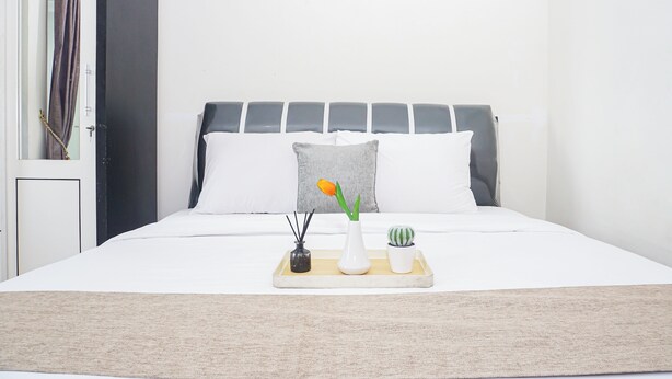 Een bed met een witte matras, een grijze hoofdeinde en witte kussens. Een dienblad met een vaas, een klein plantje en kandelaar staat op het bed.