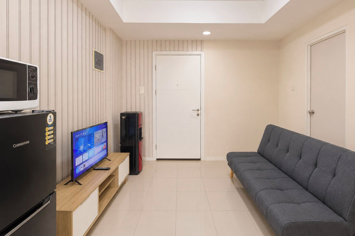 Una sala de estar moderna con un televisor de pantalla plana, un refrigerador, una computadora de escritorio y un sofá gris.