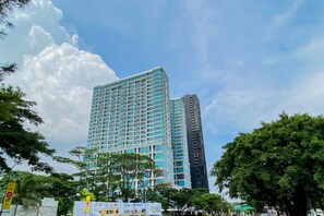 A high-rise building with a modern glass facade, surrounded by lush greenery and under a partly cloudy sky. - Decent Living In 1Br At Apartment Brooklyn Alam Sutera (Tangerang)