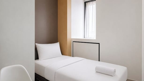 A single bed with a white mattress and pillow, a wooden headboard, a white chair, and a window with white curtains. - Good Minimalist Studio Room At Apartment Bandaraya - Tallasa City Makassar (Makassar (Ujung Pandang))